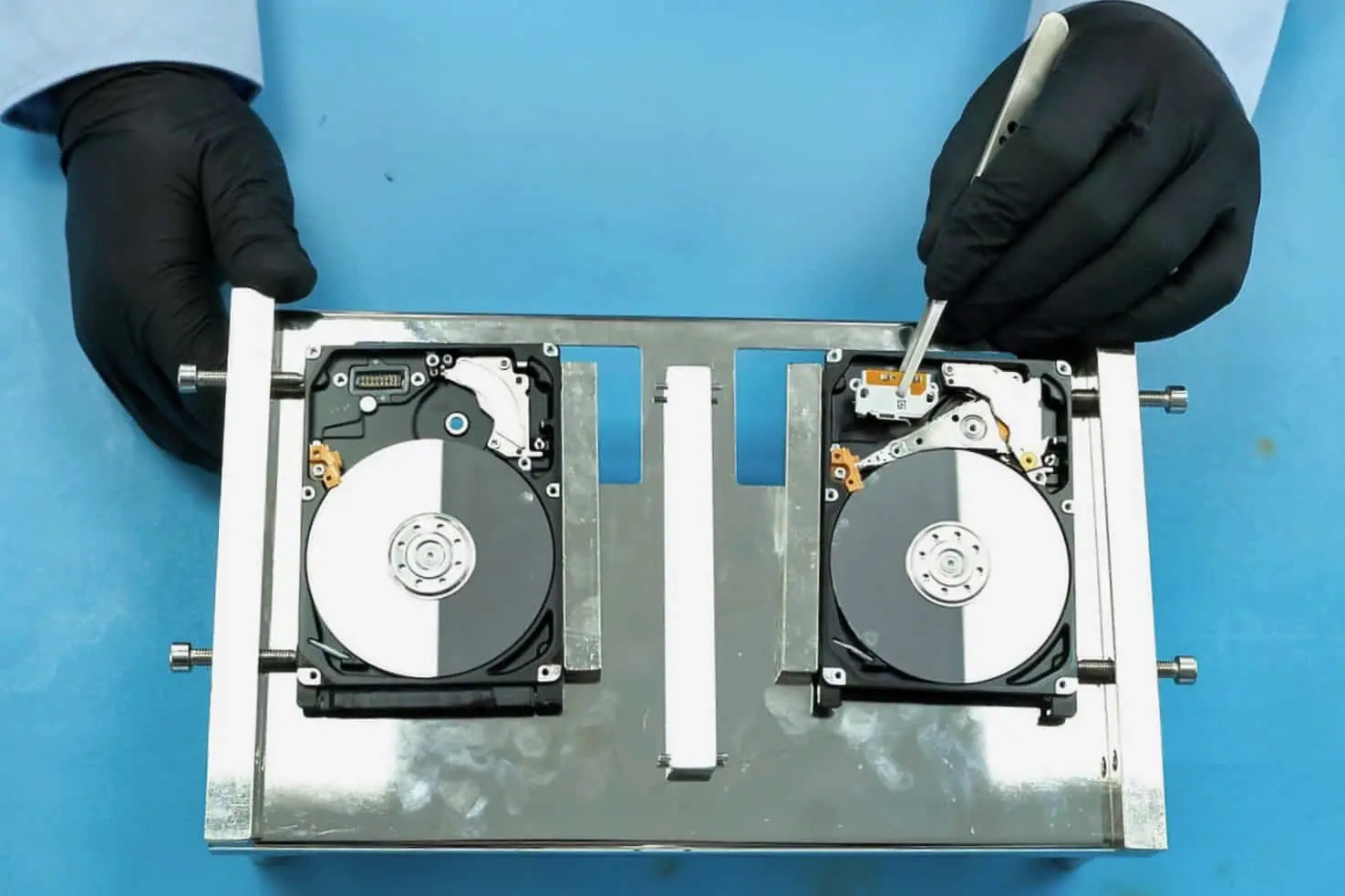 Two opened hard drives clamped in a head-swap jig, a gloved technician lifting the head assembly clear of the platter