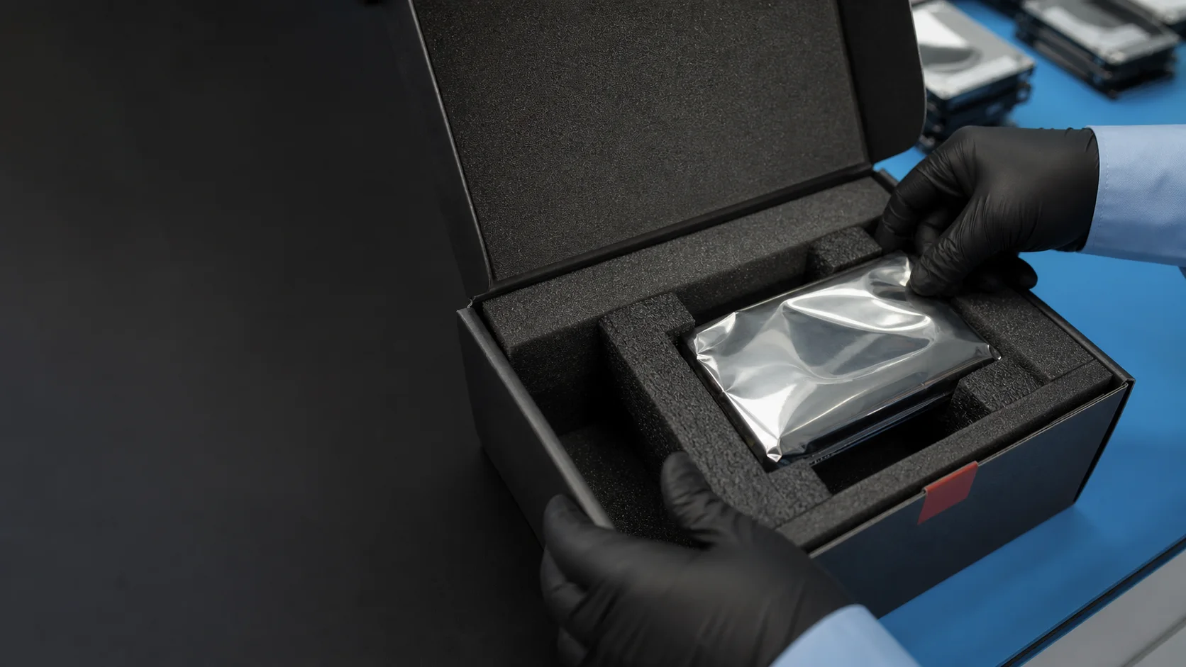 Black-gloved hands positioning a hard drive sealed in an anti-static bag inside a rigid foam-lined shipping box